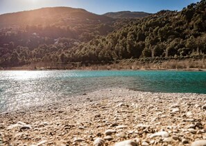 A pebble beach with clear turquoise water and forested hills in the background.