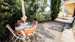 En udendørs terrasse med et glasbord og stole, omgivet af grus og grøn vegetation.
