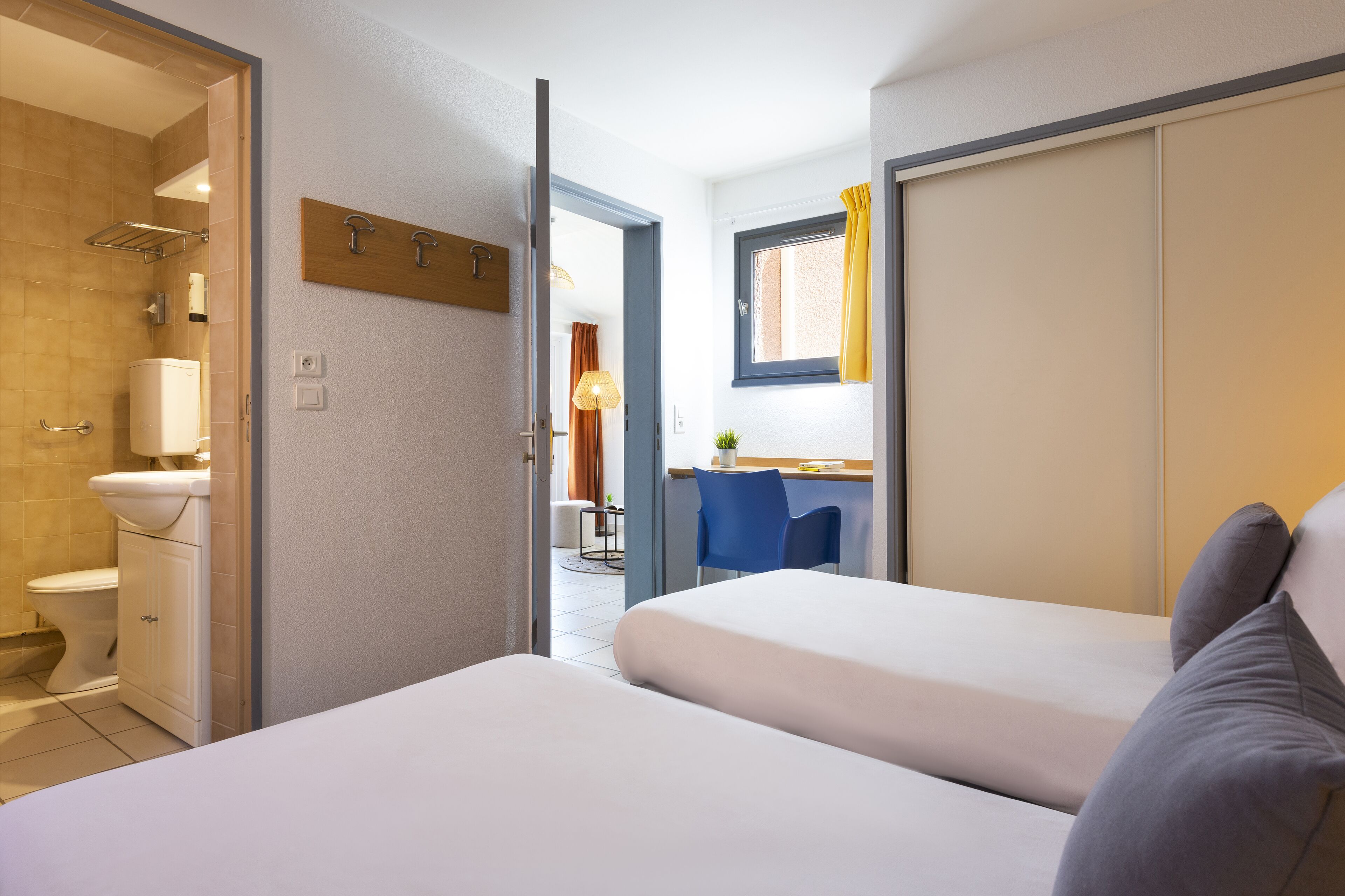 Une chambre d’hôtel avec deux lits, une salle de bain avec des toilettes et une douche, un petit bureau avec une chaise et une fenêtre avec des rideaux.