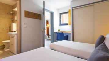 Une chambre d’hôtel avec deux lits, une salle de bain avec des toilettes et une douche, un petit bureau avec une chaise et une fenêtre avec des rideaux.