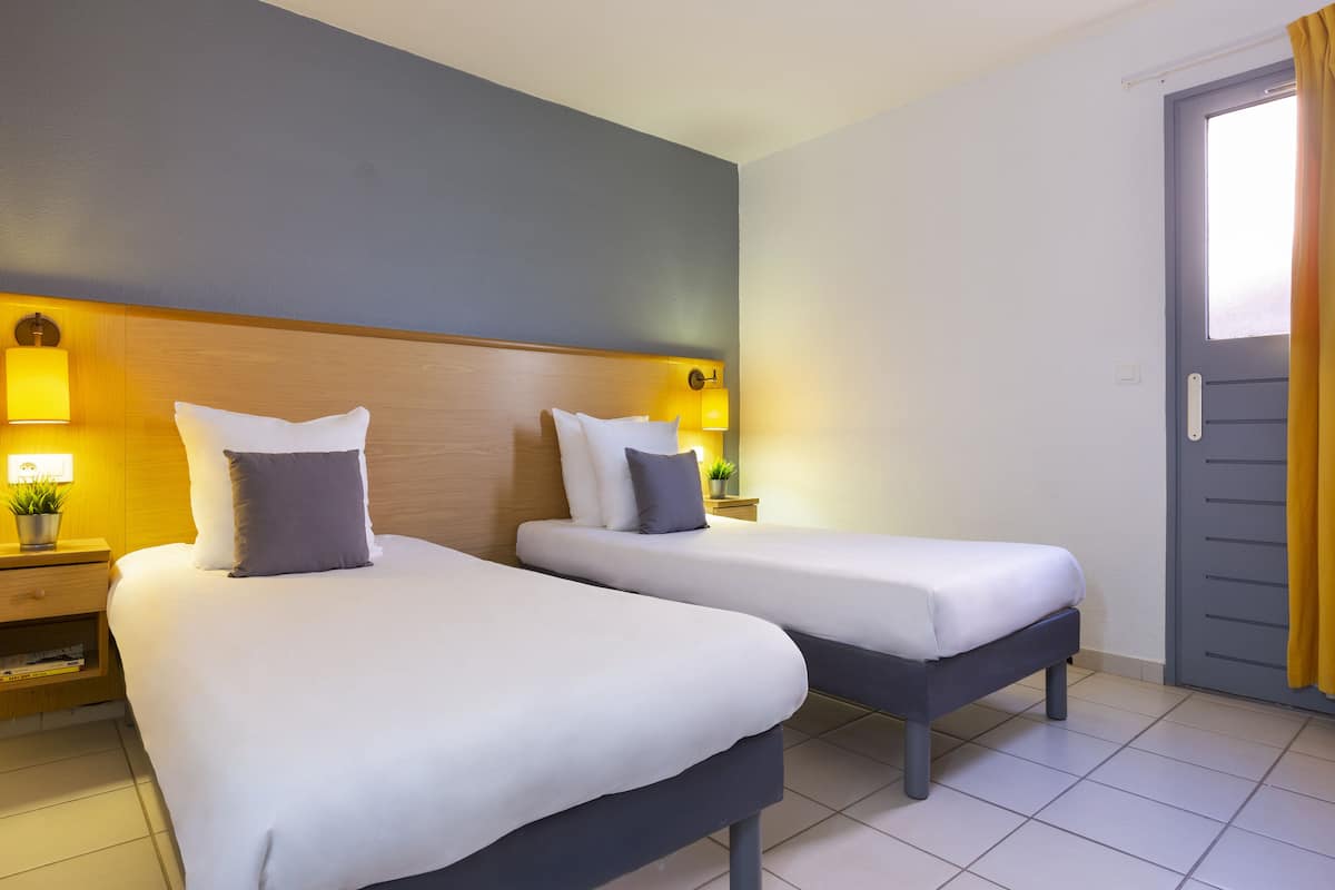 A hotel room with two beds, a wooden headboard, bedside lamps, a small table with a plant, and a door with curtains.