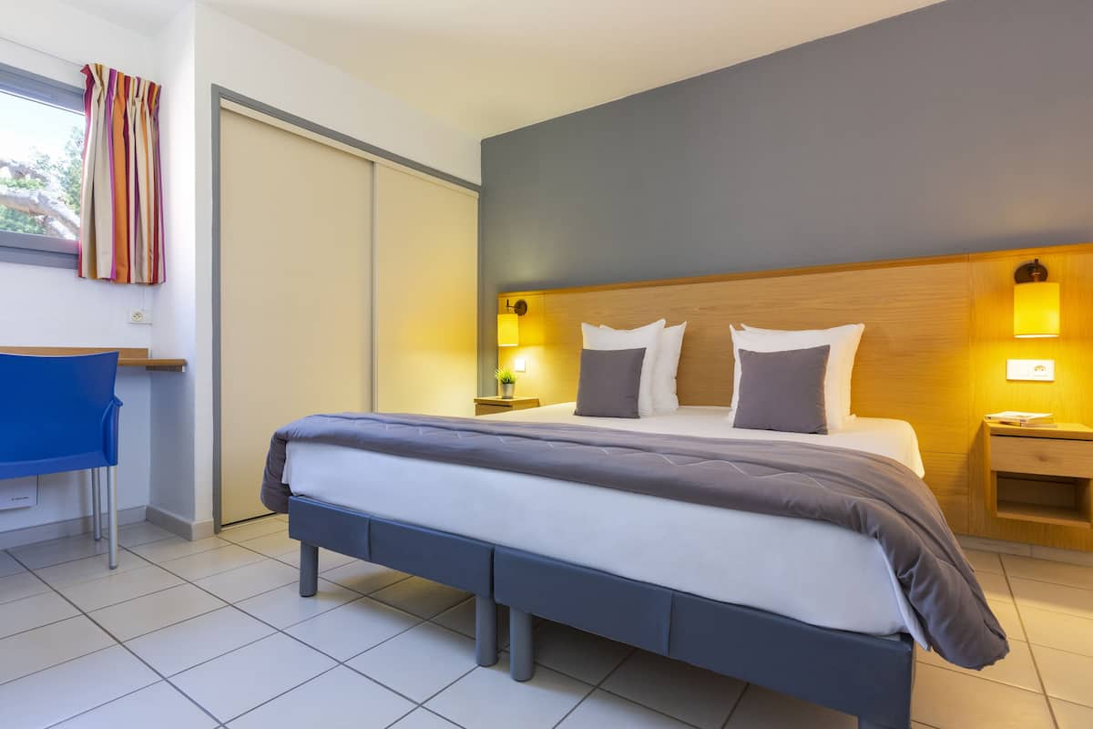 A modern hotel room with a large bed, bedside tables with lamps, a desk with a chair, and a view of the outdoors through a window.