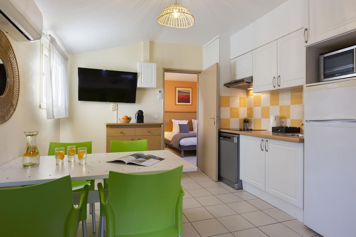 A compact kitchen with white cabinets, a microwave, and a sink. There is a dining area with a table and chairs, and a television mounted on the wall.