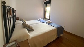 A single bed with a wooden headboard, a window with blinds, and a small bedside table with a lamp.