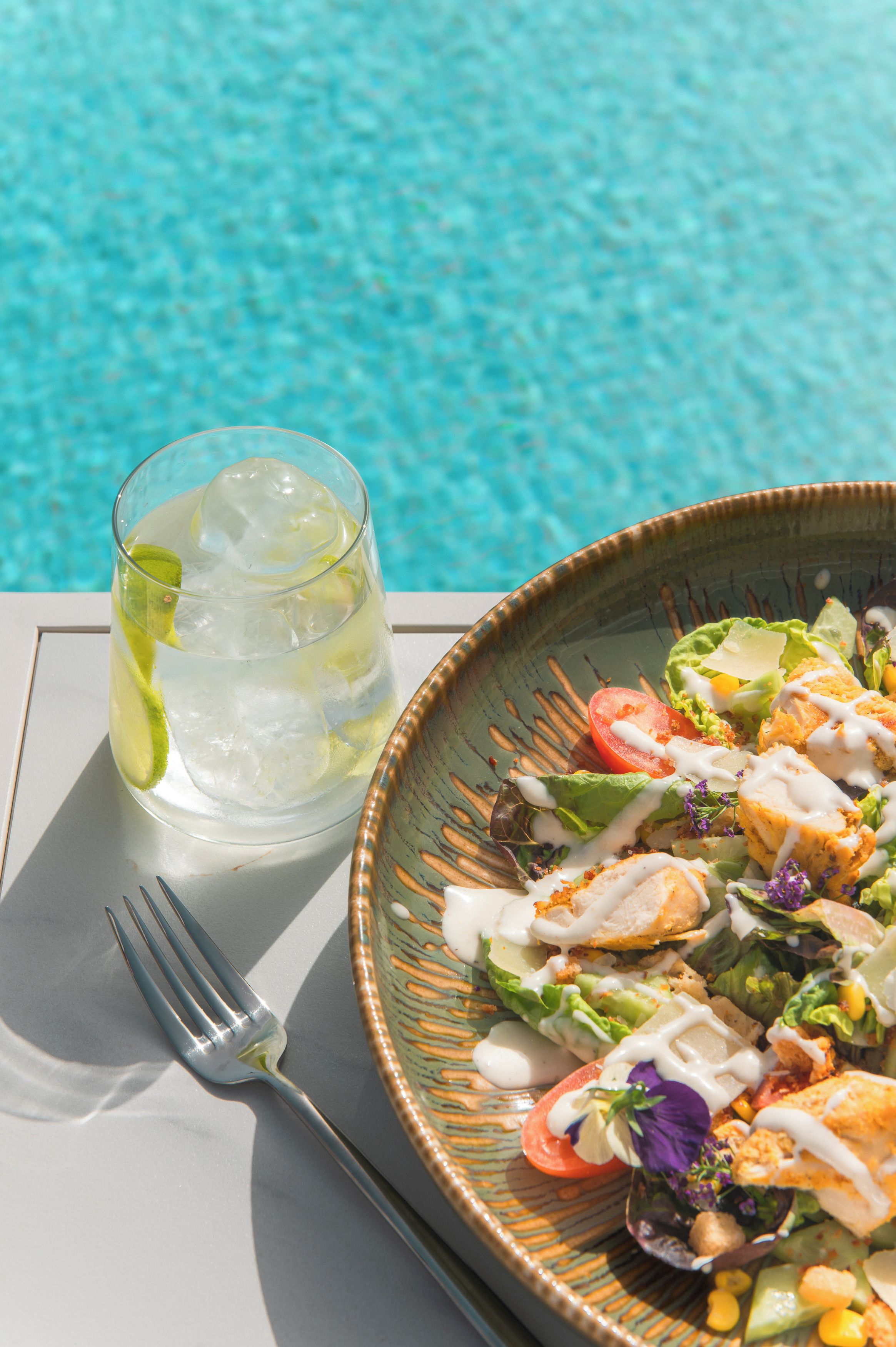 Spisebord med mad og et glas isdrik ved en pool.