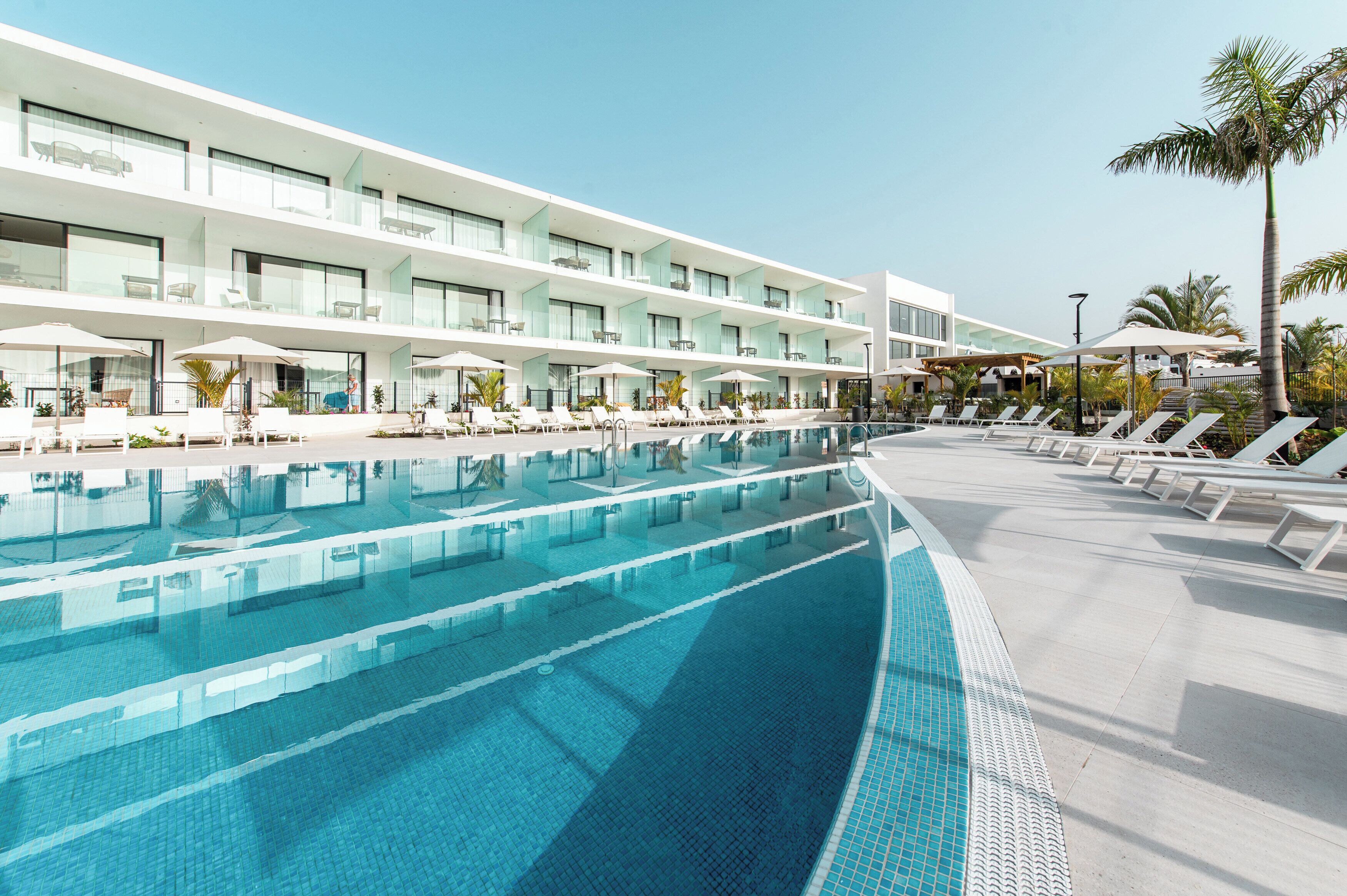 A modern hotel with a large pool, sun loungers, and a clear blue sky.