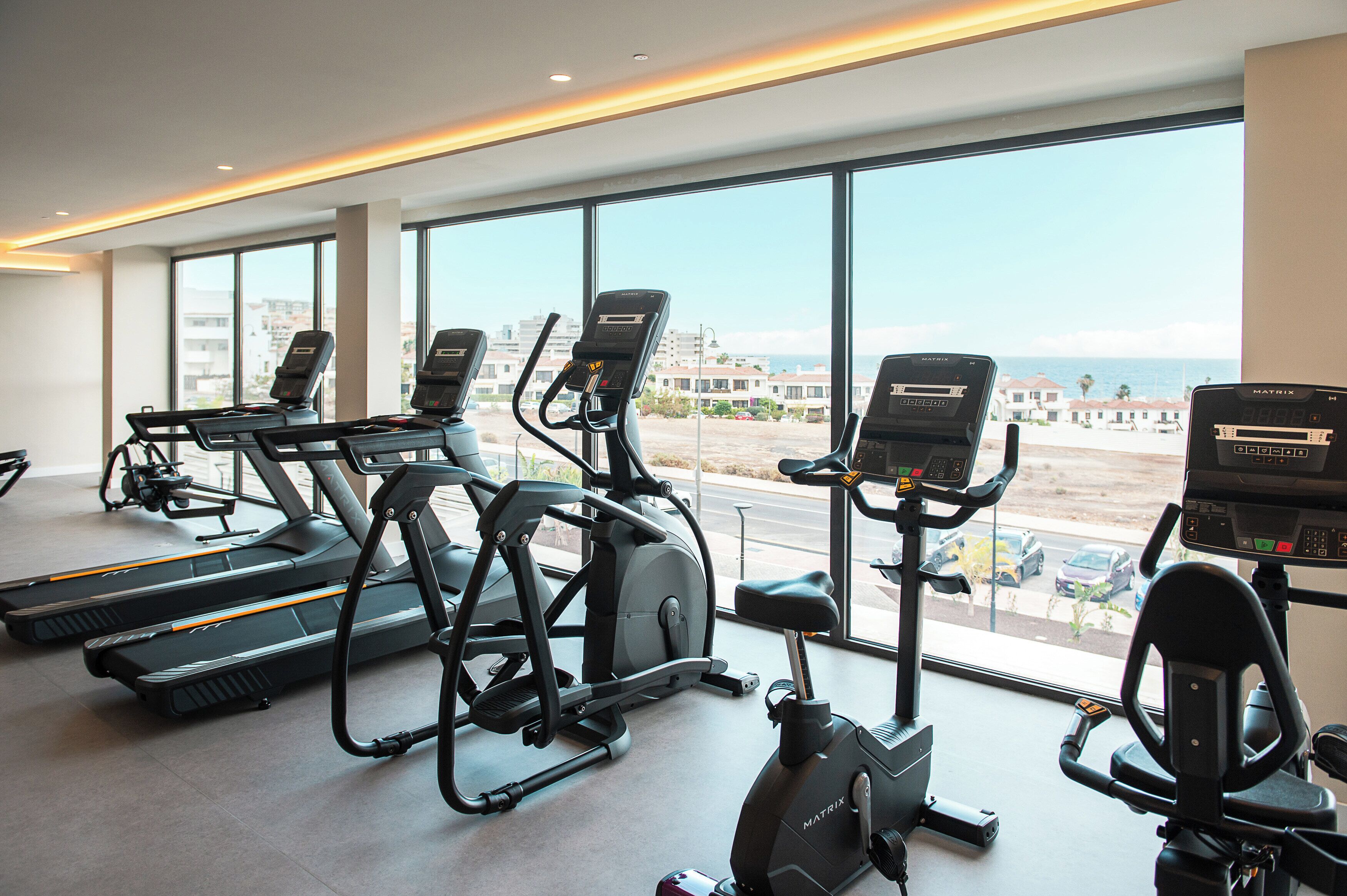 A gym with treadmills, elliptical machines, and stationary bikes, large windows with a view of buildings and a road.