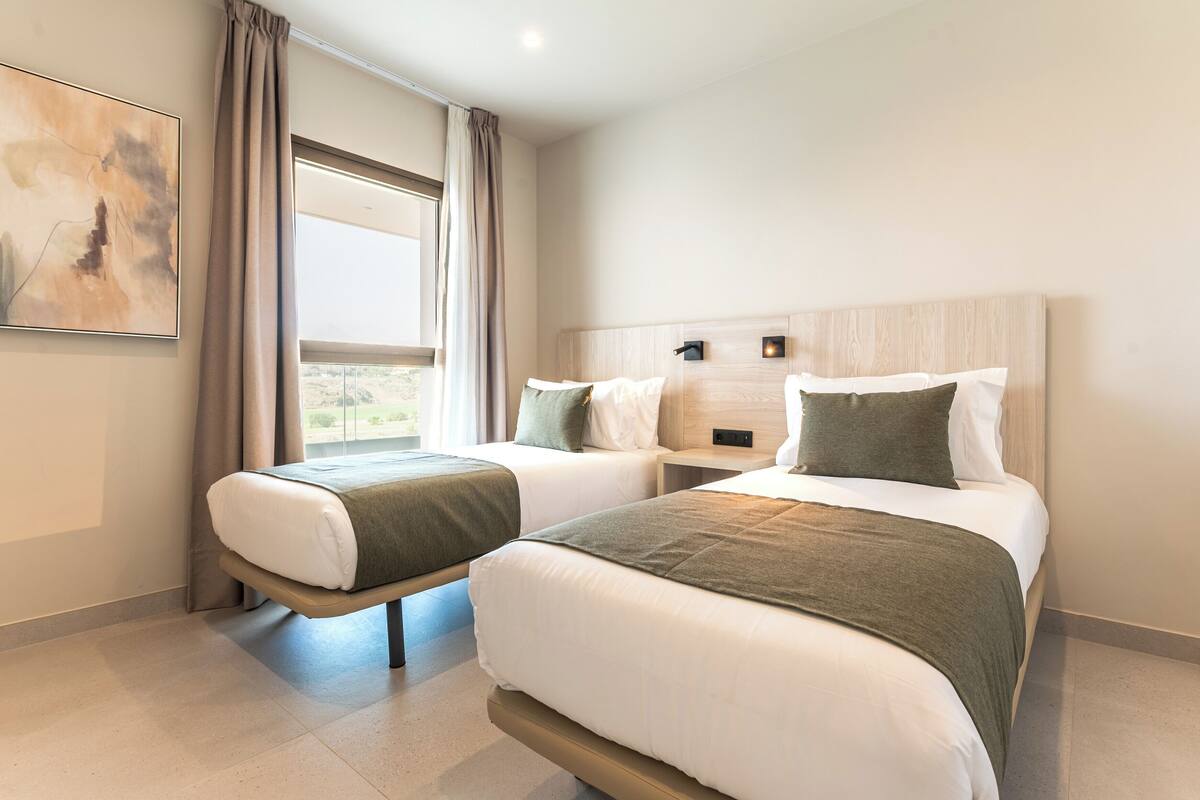 Une chambre d’hôtel avec deux lits, une grande fenêtre avec des rideaux, une œuvre d’art accrochée au mur et un plancher de couleur claire.