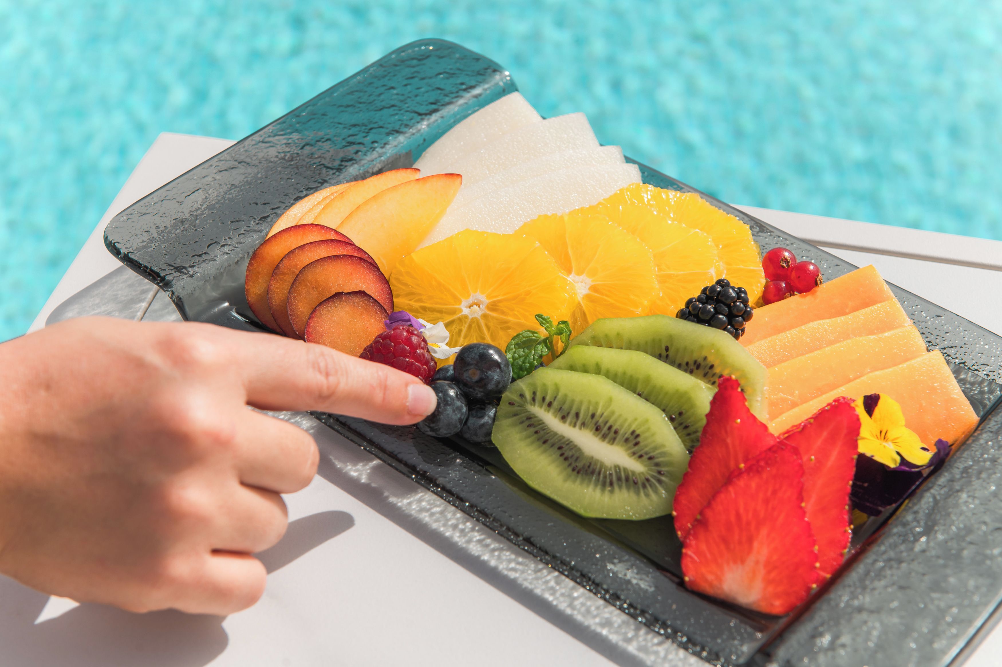 A fruit platter with various fruits and berries on a table by a pool.