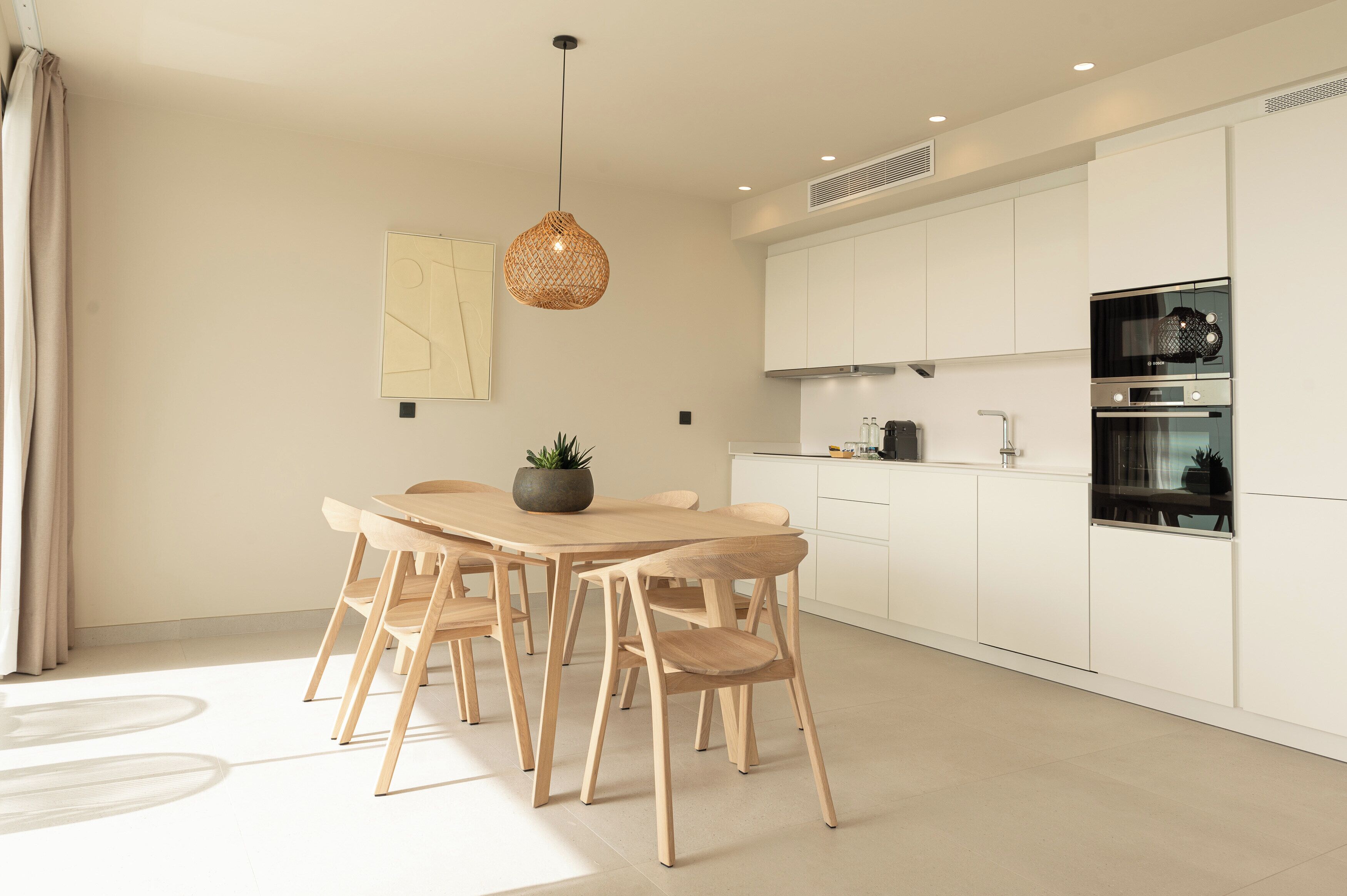 Una cocina moderna con armarios blancos, una mesa de comedor con sillas, una lámpara colgante y una planta en maceta.