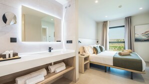 A modern hotel room with a large mirror, a wooden vanity, a bed with a view, and a small bedside table.
