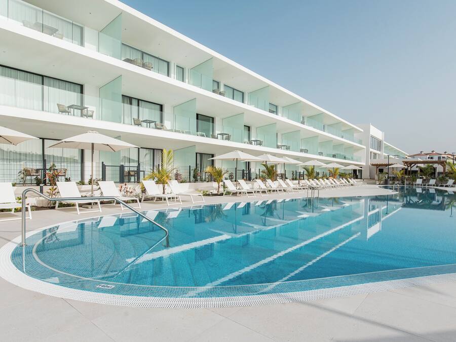 A modern hotel with a large pool, sun loungers, and a clear sky.