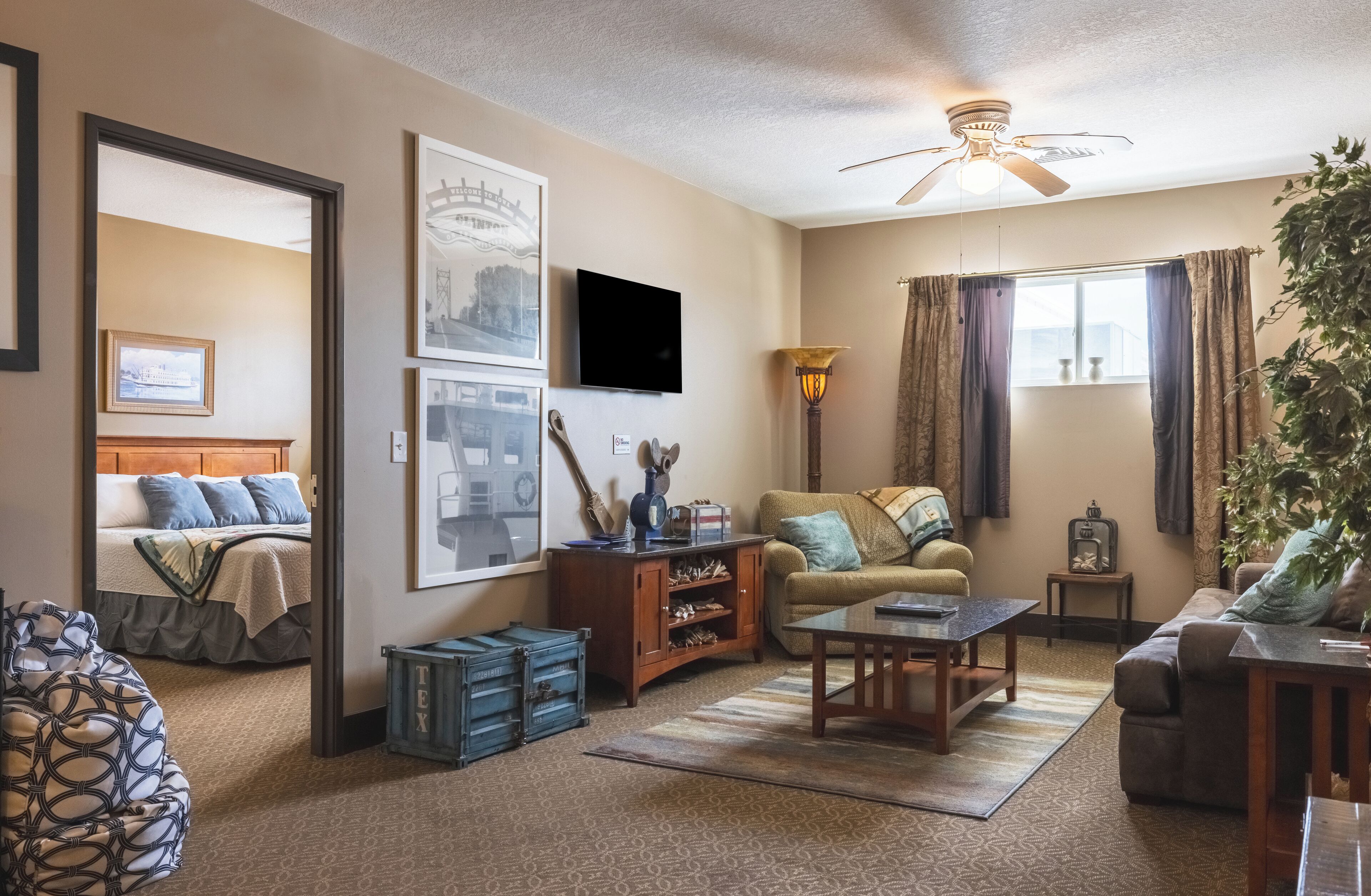A living room with a sofa, coffee table, and a TV. There is a bed with a headboard and a window with curtains.