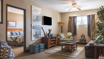 A living room with a sofa, coffee table, and a TV. There is a bed with a headboard and a window with curtains.