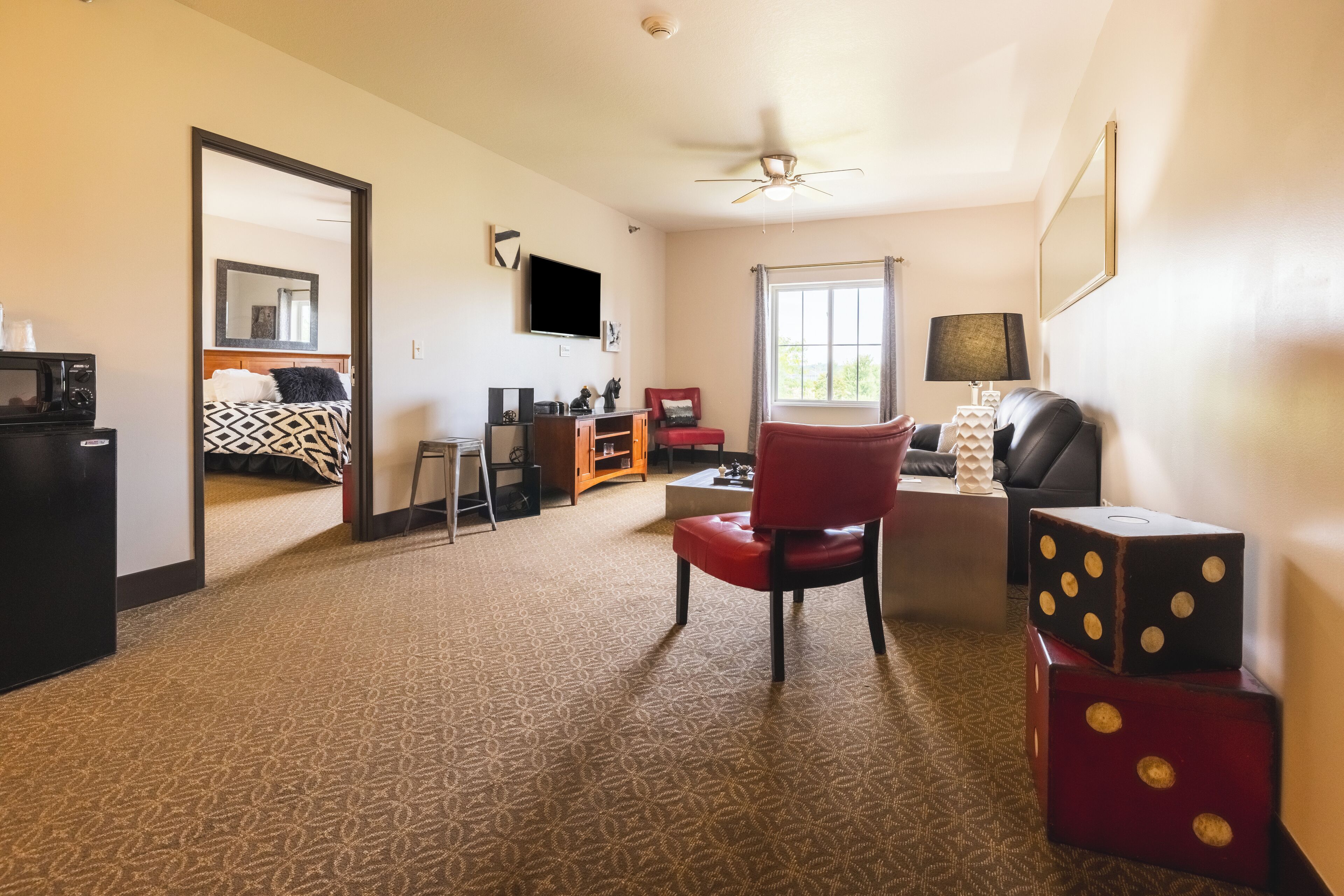 Habitación de hotel con alfombra estampada, una silla roja, televisión, ventilador de techo y una ventana con cortinas.
