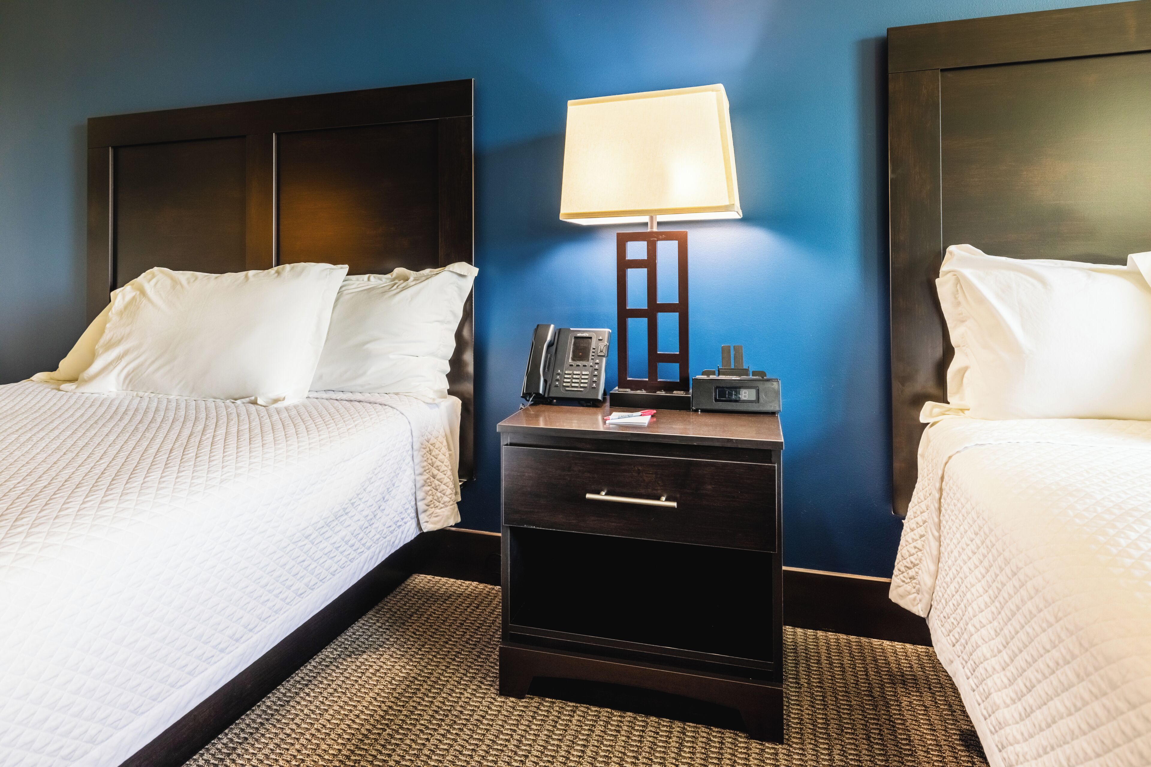 A hotel room with two beds, a nightstand with a lamp, a phone, and a drawer.