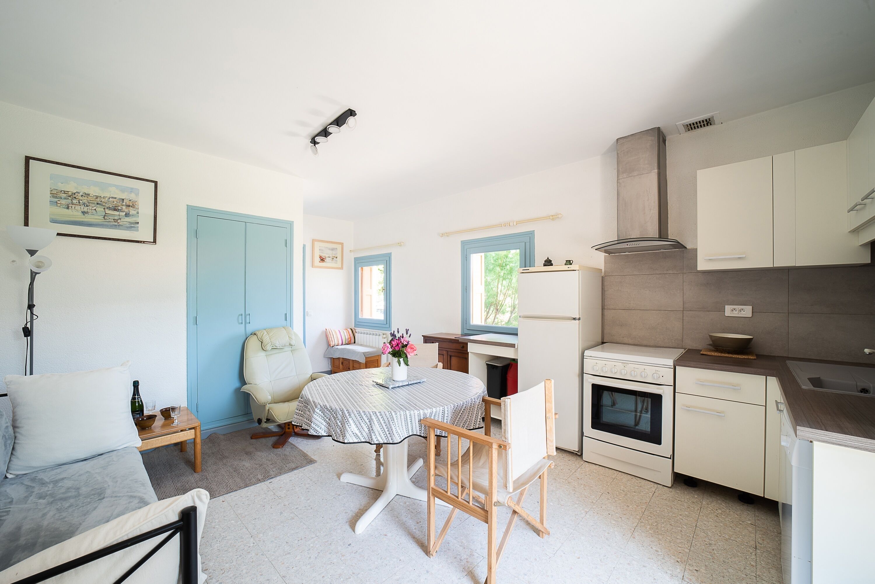 Une cuisine compacte comprenant un coin repas, un canapé et une chaise. On y trouve une table avec un vase de fleurs, une fenêtre aux cadres bleus et une cuisinière.