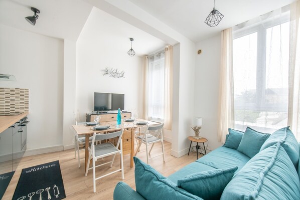A modern living room with a teal sofa, a dining table with chairs, a TV, and a kitchen area with wooden cabinets. - La Pause Marine - Charmant appt pour 4 (Calais)