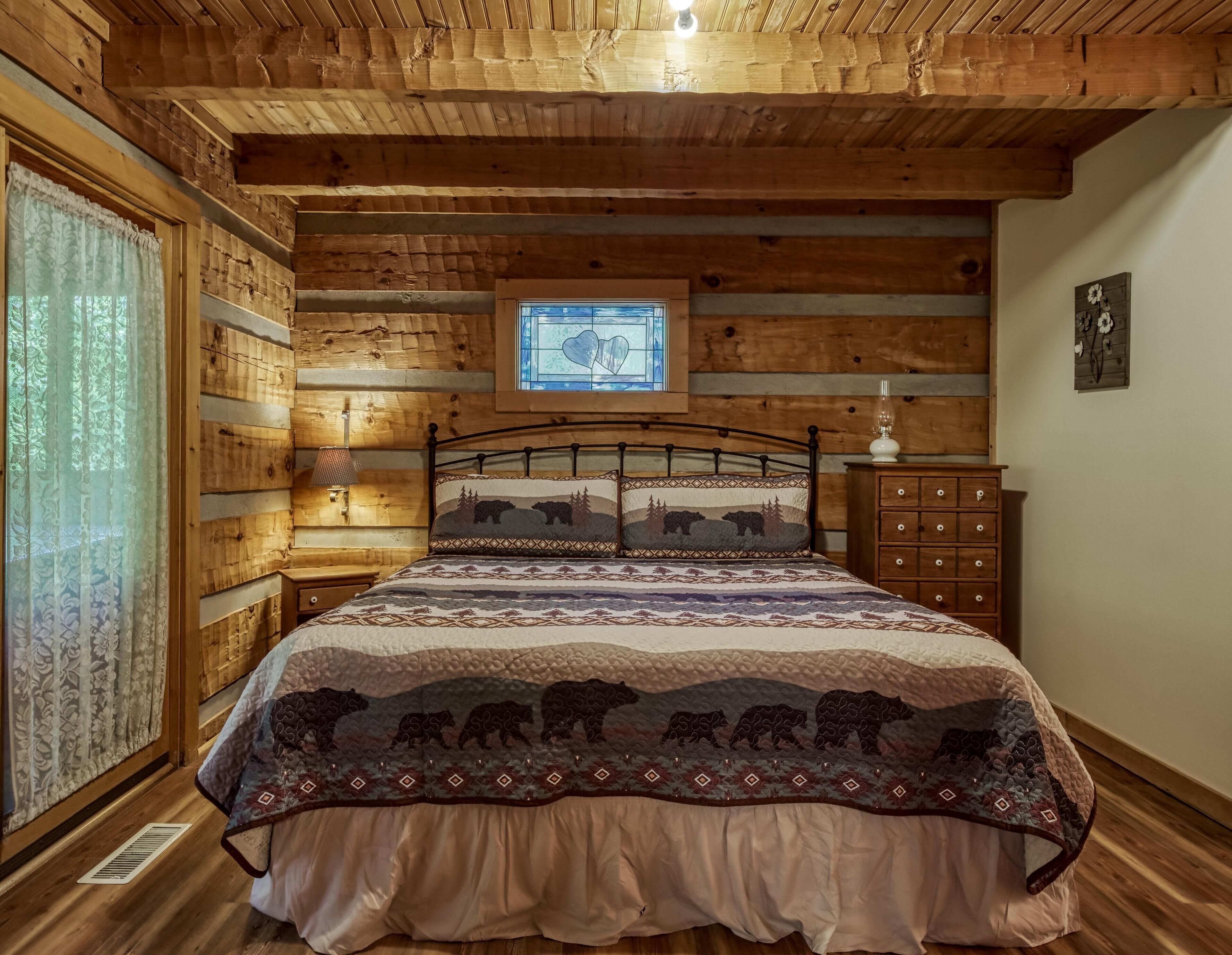 Un dormitorio de estilo cabaña de madera con una cama, una mesita de noche, un tocador y una ventana con un vitral en forma de corazón.