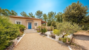 Une maison avec une allée en gravier menant à une porte bleue, entourée de verdure et d’arbres.