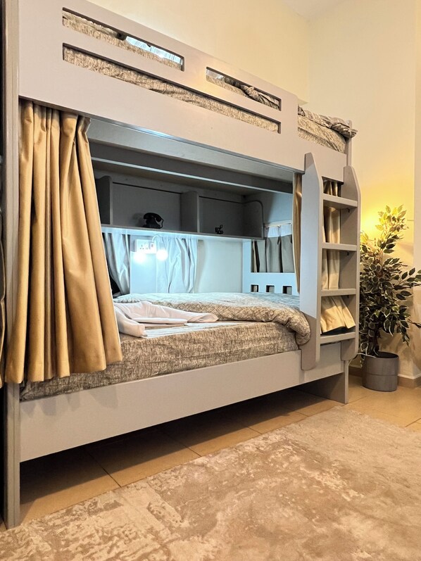 Un lit superposé avec un bureau et une étagère, une plante et un rideau.