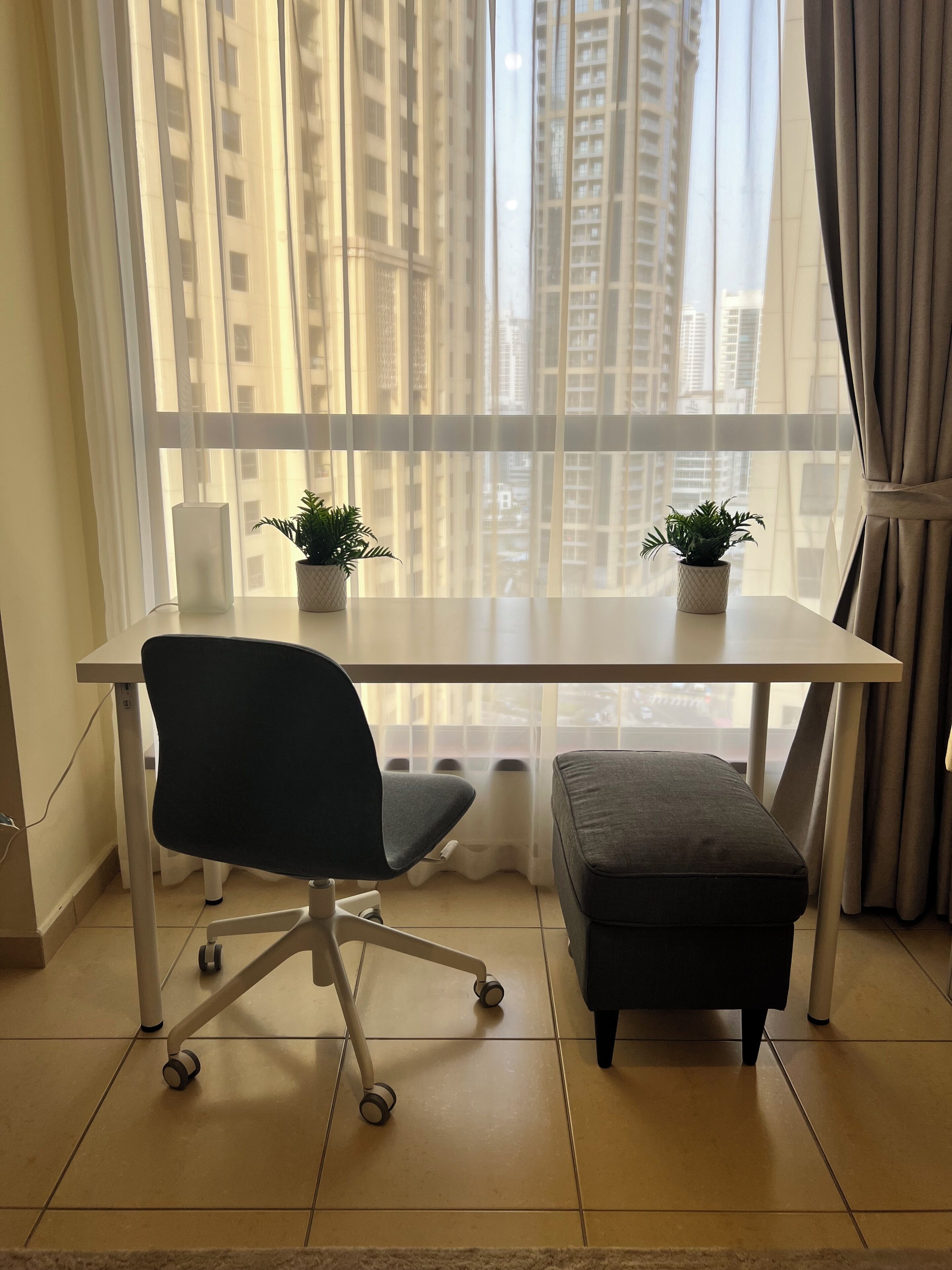A modern office space with a white desk, black office chair, and an ottoman. There are two potted plants on the desk.