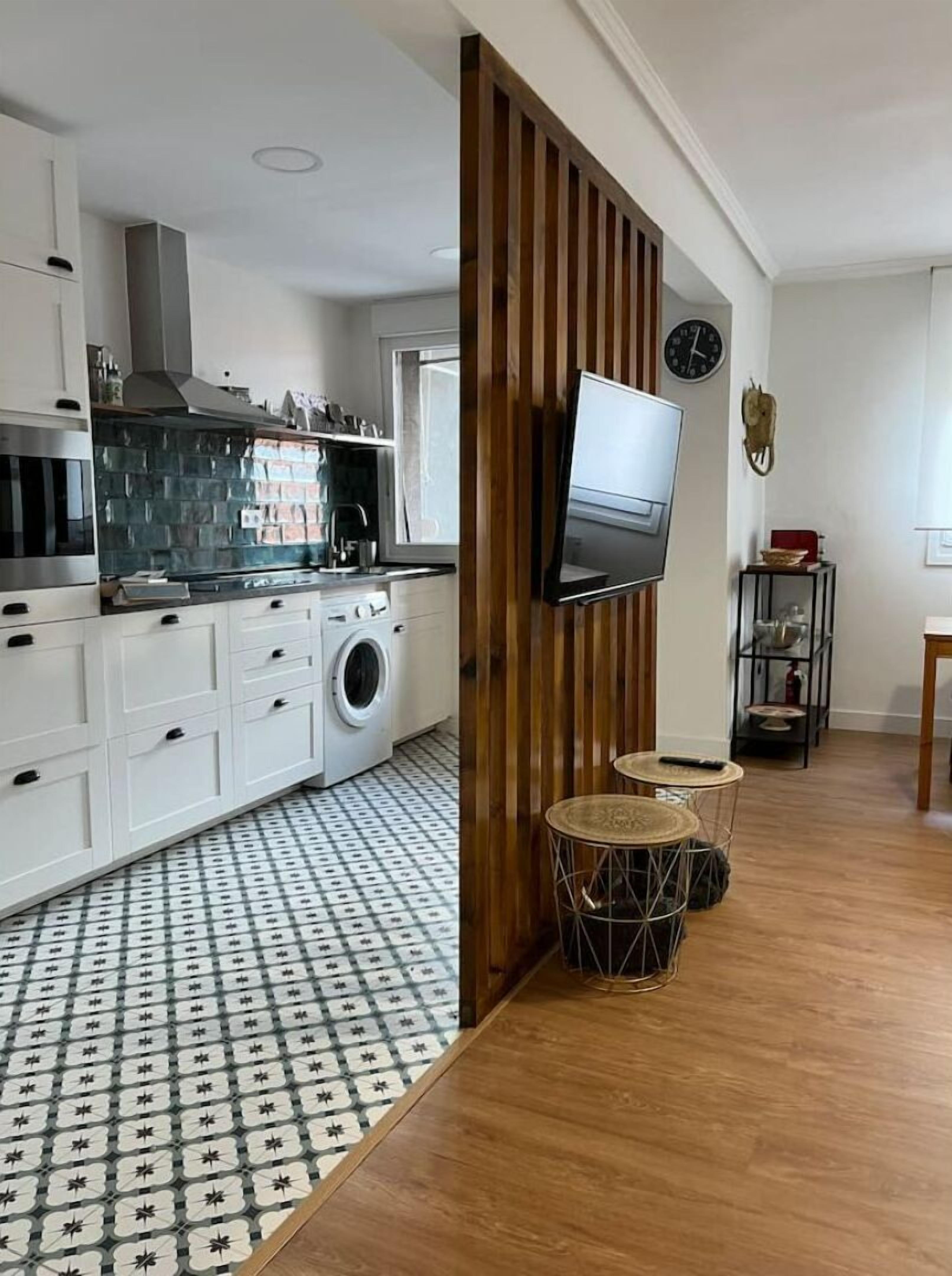 Une cuisine moderne avec des meubles blancs, une cuisinière encastrée et un lave-linge. Un séparateur en bois est installé, et un téléviseur y est fixé. Le sol est recouvert de carreaux décoratifs formant un motif.