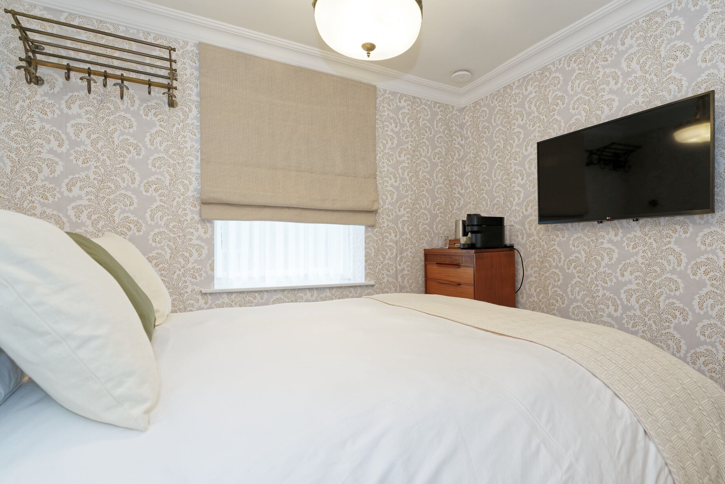 A bedroom with a bed, a TV mounted on the wall, a window with a blind, and a wooden dresser.