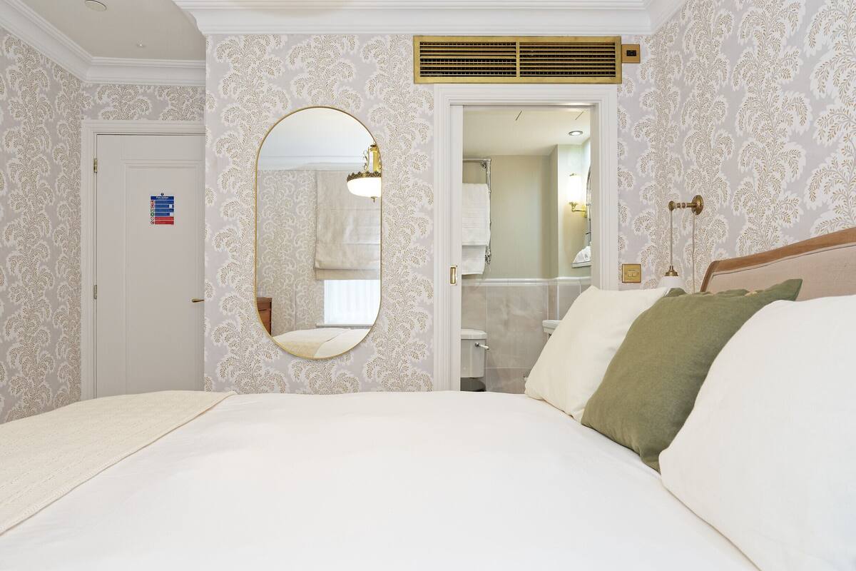 A hotel room with a large bed, a round mirror, a door with a towel rack, and a bathroom visible through an open door.