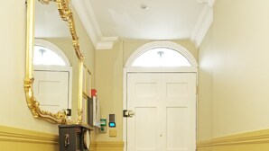 A hallway with a tiled floor, a large mirror, a door with a brass handle, and a small pedestal table.