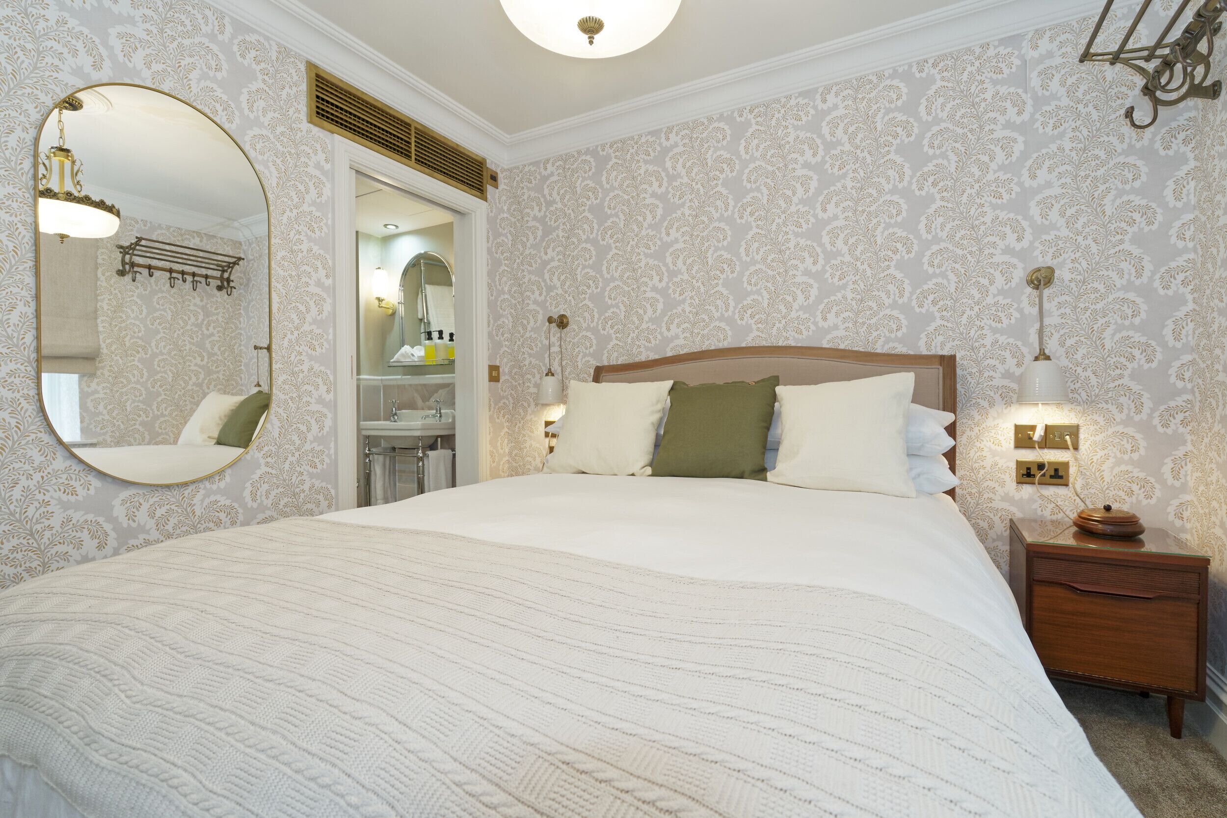 A bedroom with a large bed, a wooden nightstand, a mirror, and a wall-mounted light fixture.