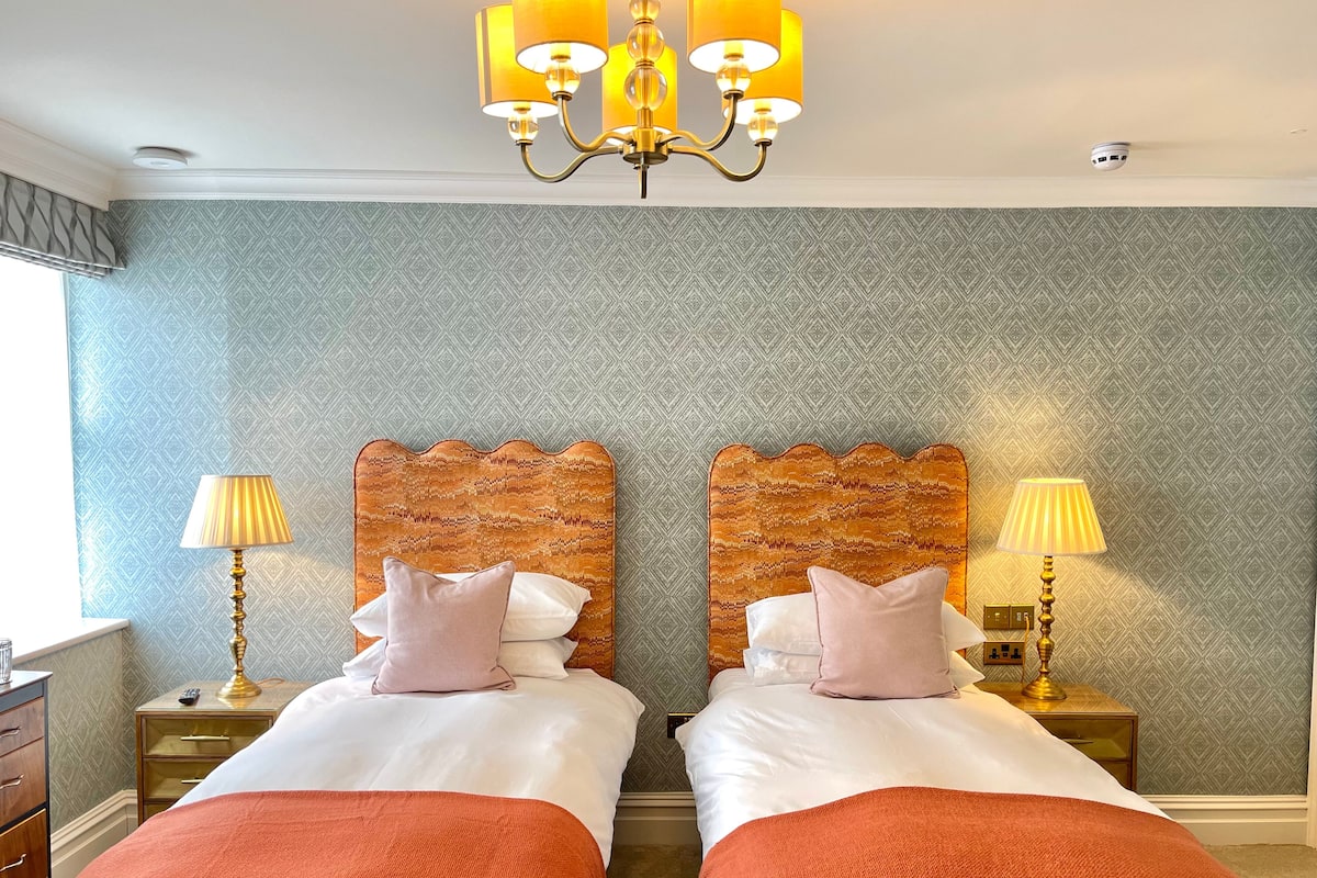 A hotel room with two beds, a chandelier, patterned wallpaper, and a window with curtains.