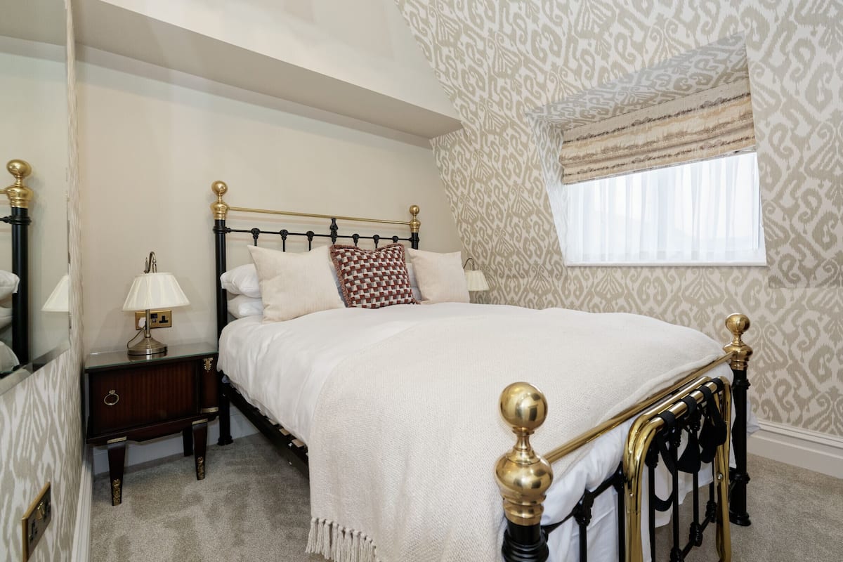 A neatly made bed with a gold headboard, a bedside table with a lamp, and a patterned wall.