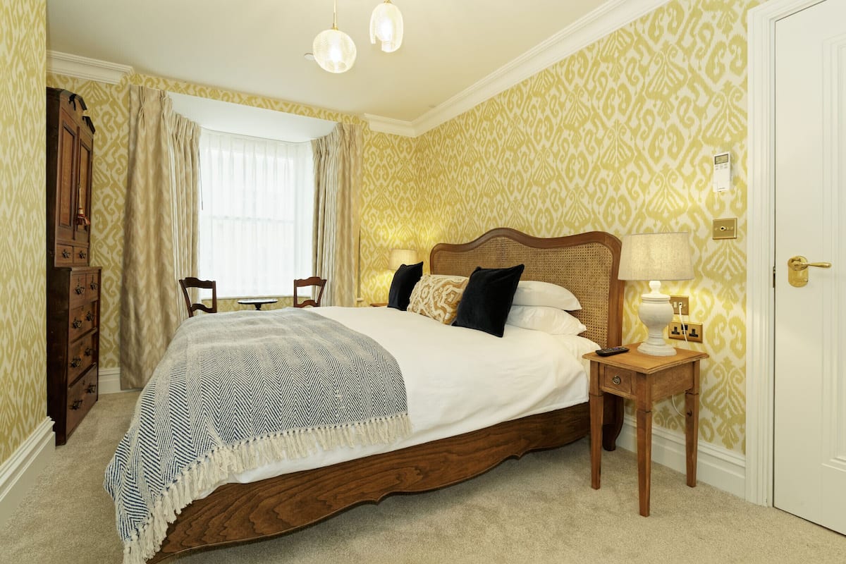 A bedroom with a large bed, a bedside table with a lamp, a dresser, and a window with curtains.