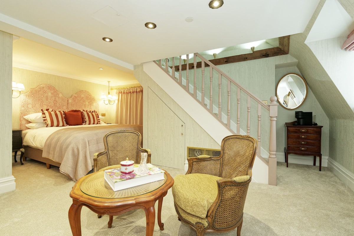 A bedroom with a bed, two wicker chairs, a small round table, a dresser, and a staircase.