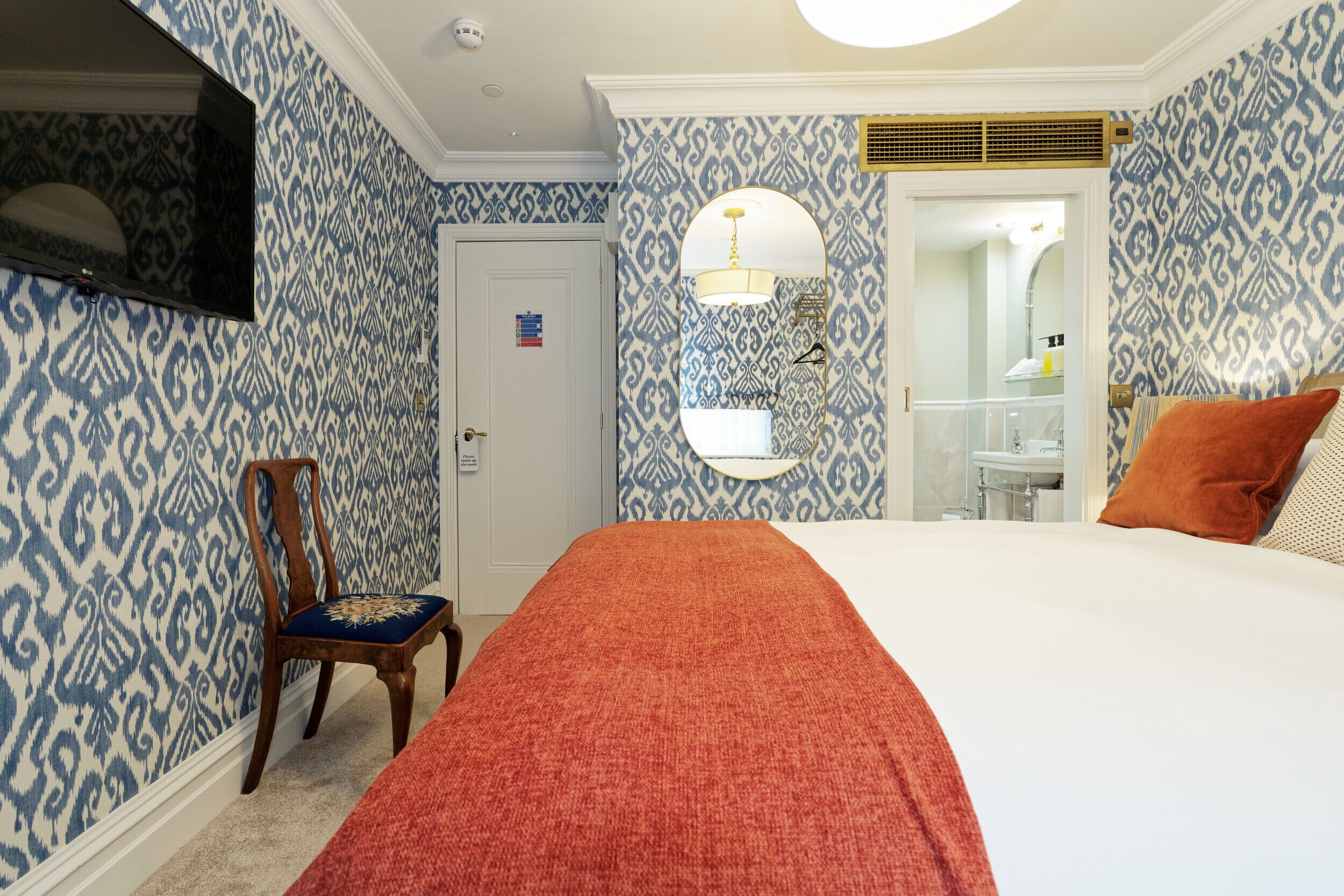 A bedroom with patterned wallpaper, a bed with a red blanket, a wooden chair, a mirror, and a bathroom visible through an open door.