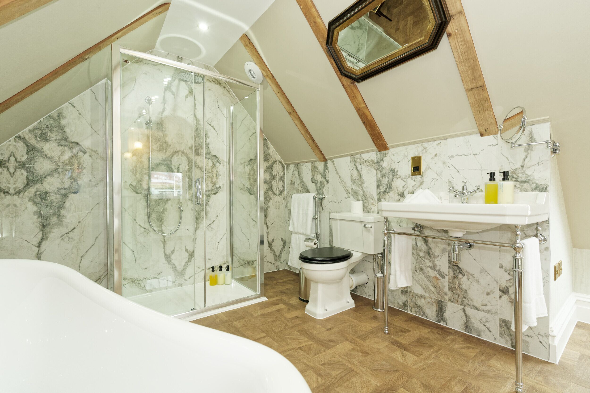 A bathroom with a glass shower enclosure, a white toilet, a sink with a marble countertop, and wooden flooring.