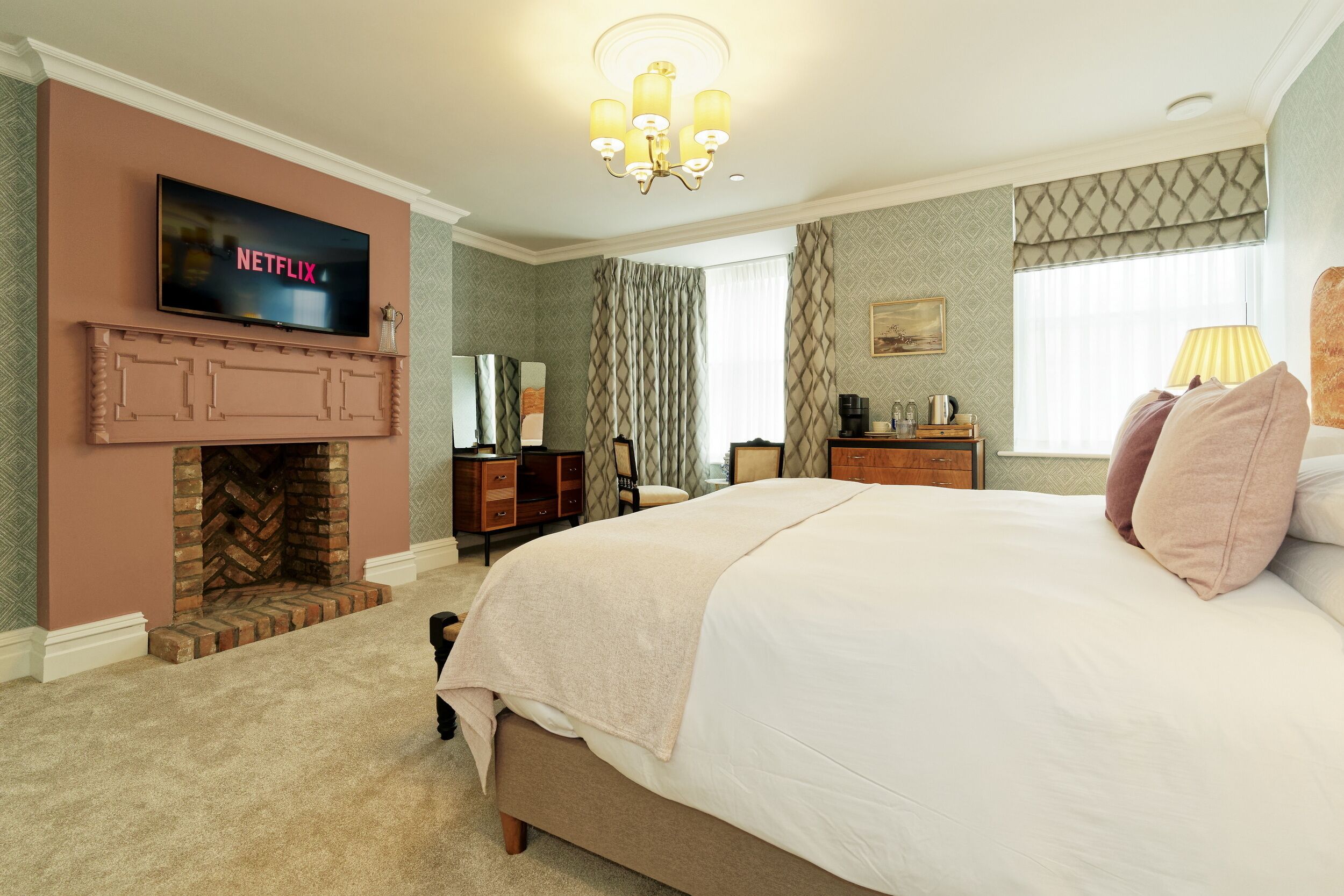 A hotel room with a large bed, a fireplace, a television mounted above it, and a desk with a lamp.