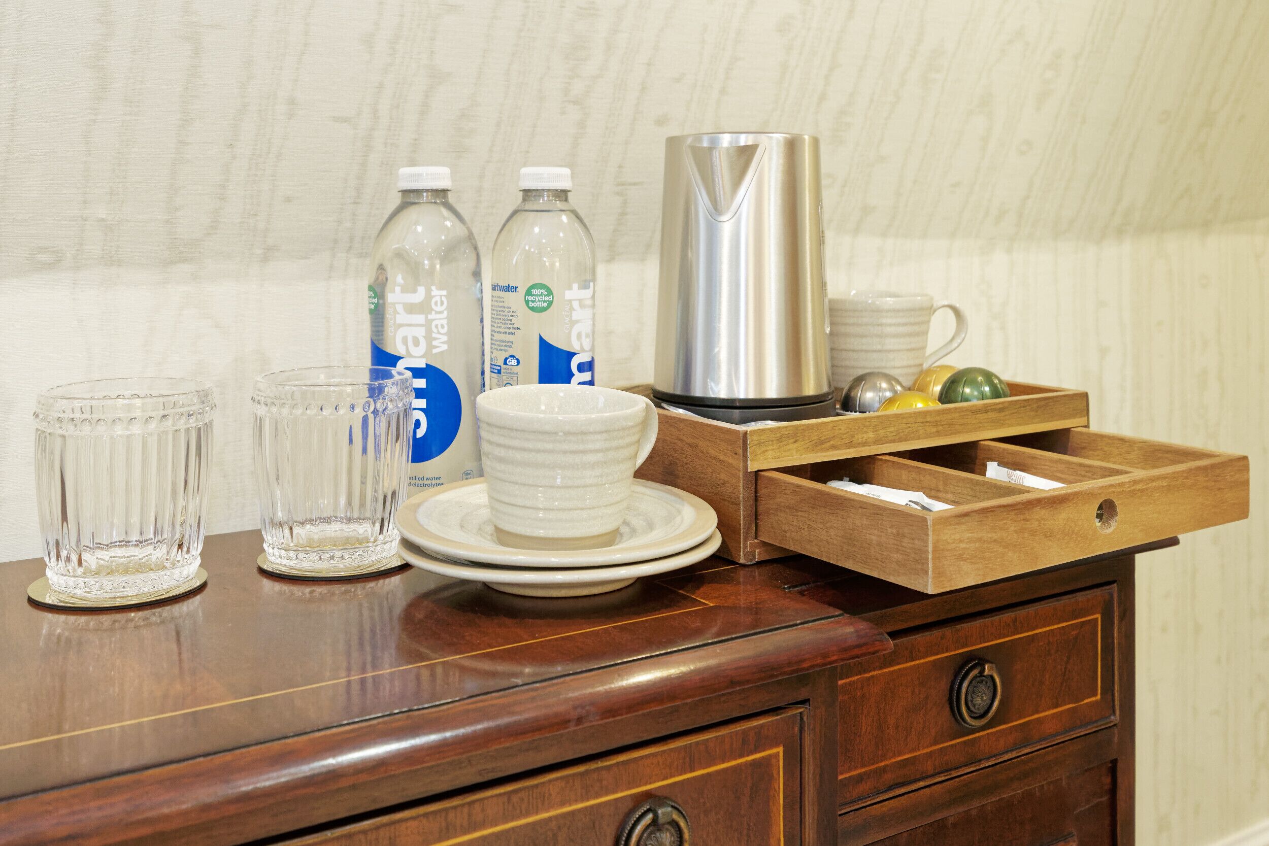 A wooden table with clear glasses, water bottles, a white mug, a stainless steel kettle, and a wooden drawer with compartments.