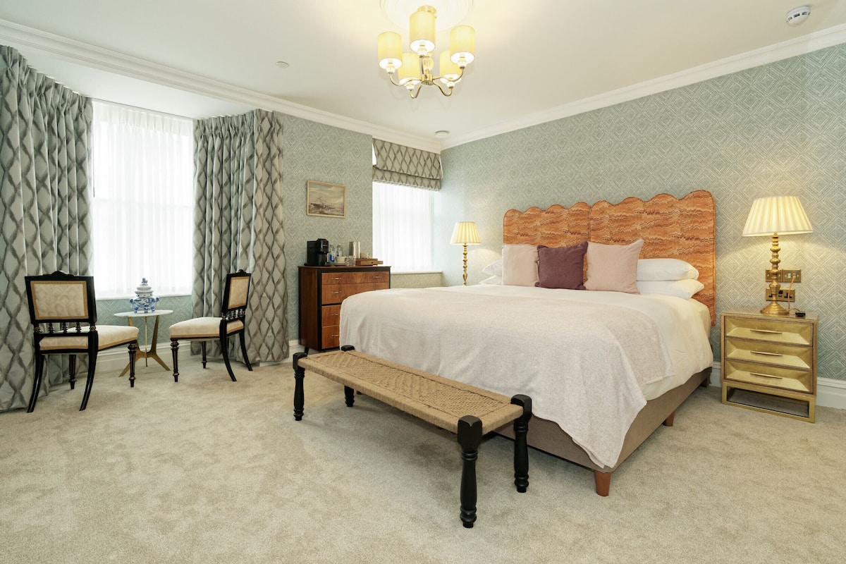 A neatly arranged bedroom with a large bed, bedside lamps, a small table with chairs, and a bench.