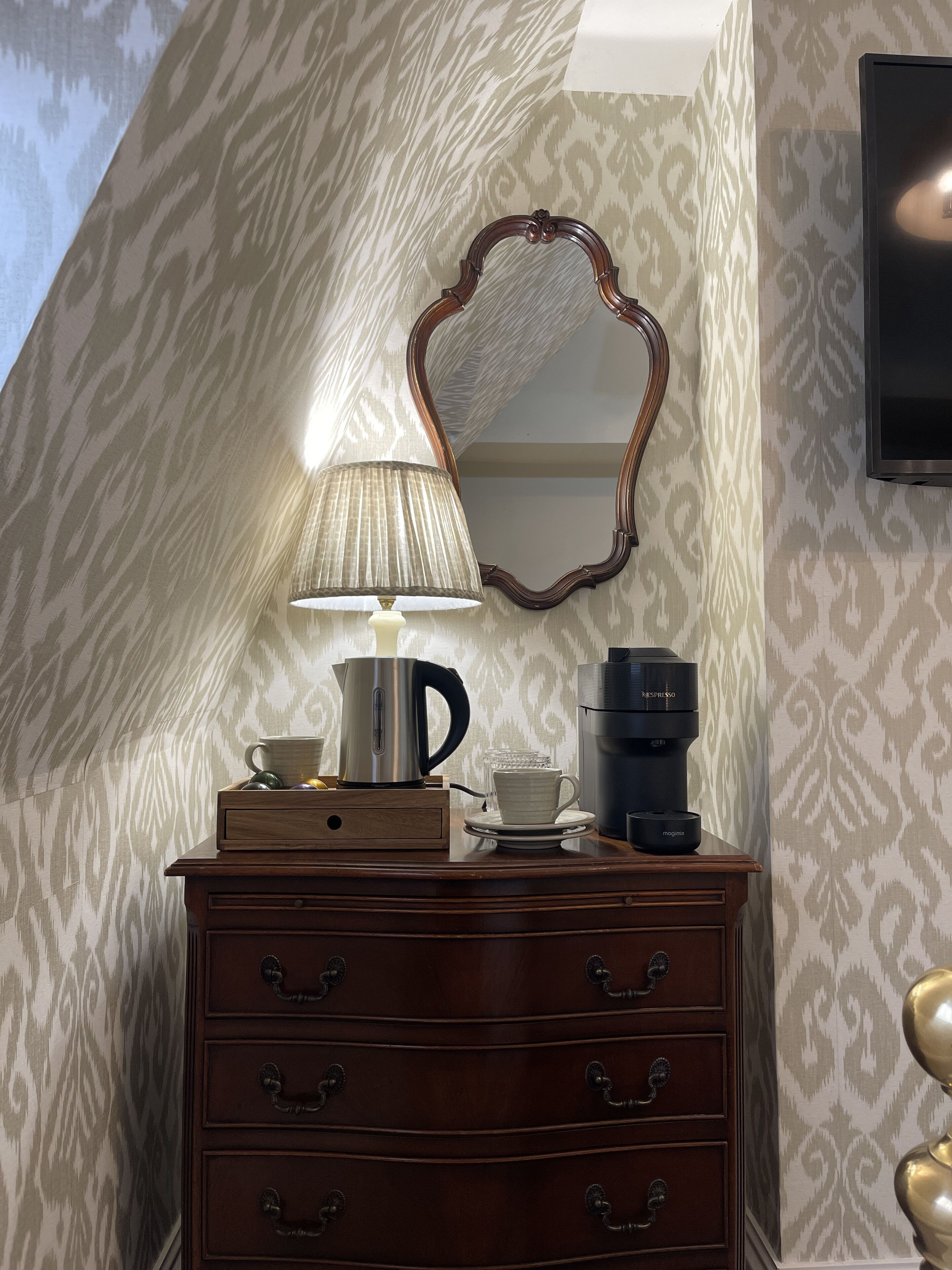Een houten dressoir met een lamp, een koffiezetapparaat en een spiegel in een kamer met behangpapier.