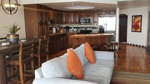 Een moderne keuken met houten kastjes, een centraal eiland en een zithoek met een witte bank en oranje kussens.