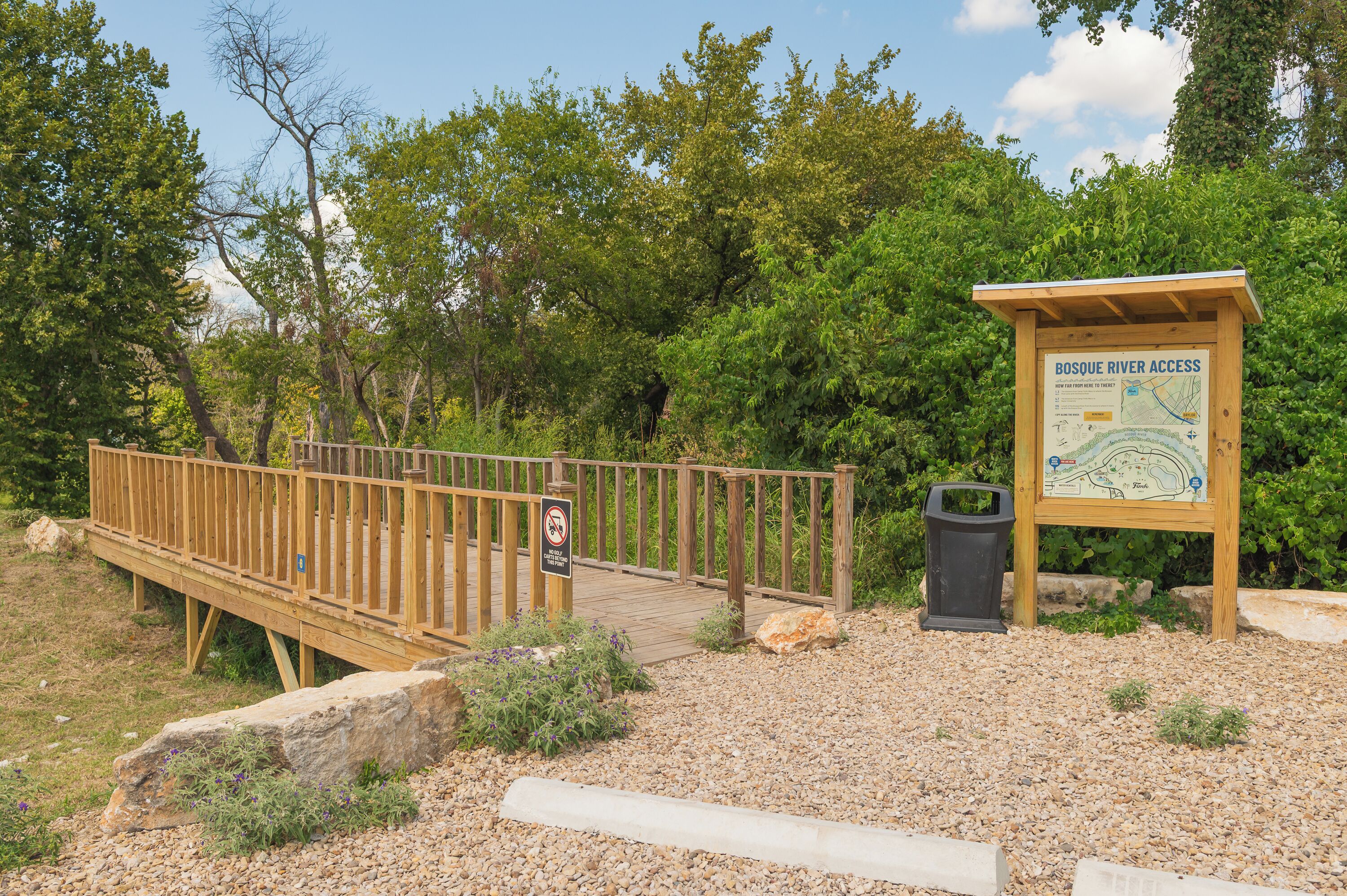 A wooden bridge with a signboard indicating Bosque River access, surrounded by greenery and a gravel path.