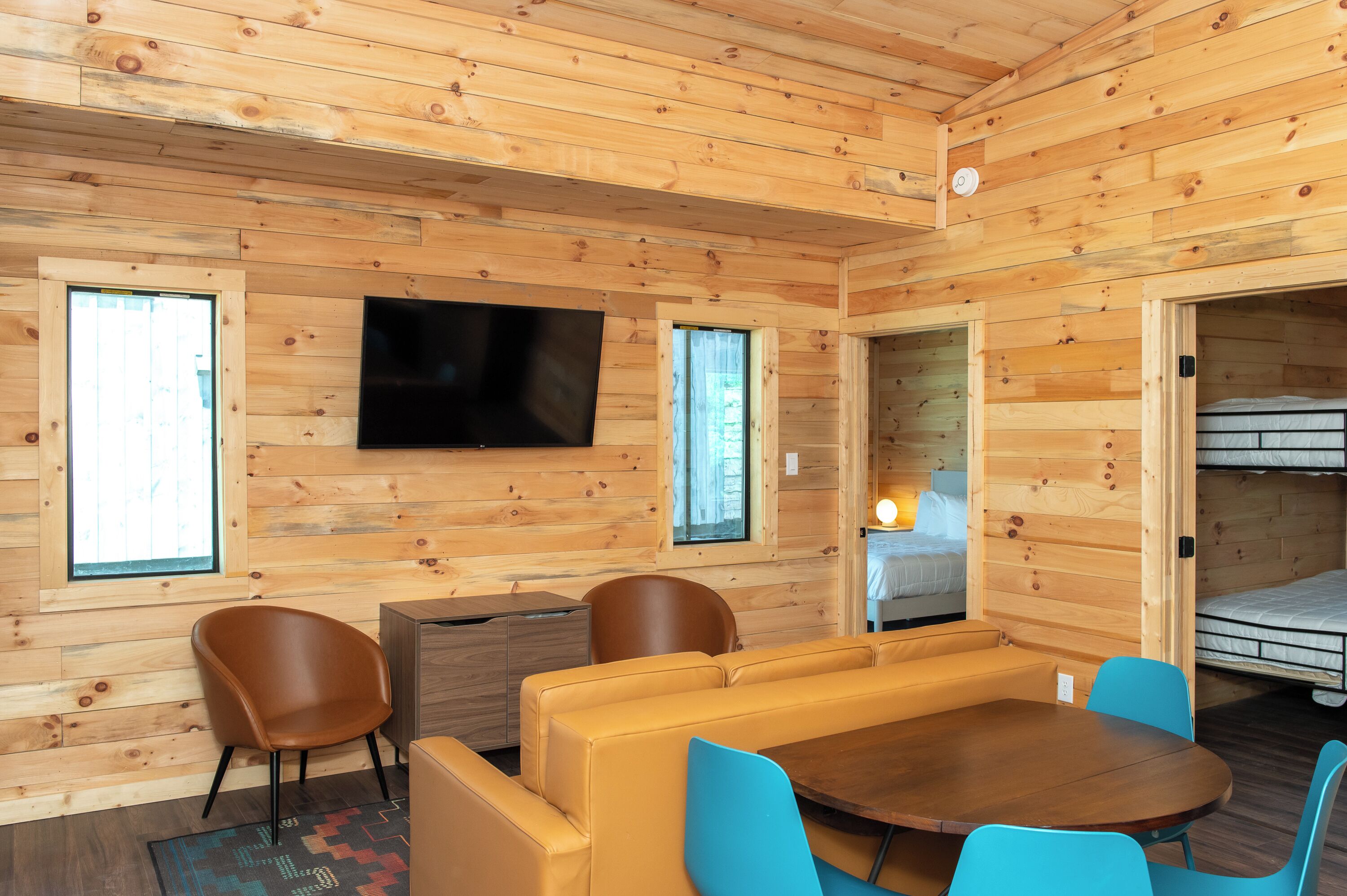 L’intérieur d’un chalet en bois, avec un canapé, des fauteuils, une table, un téléviseur et un lit superposé.