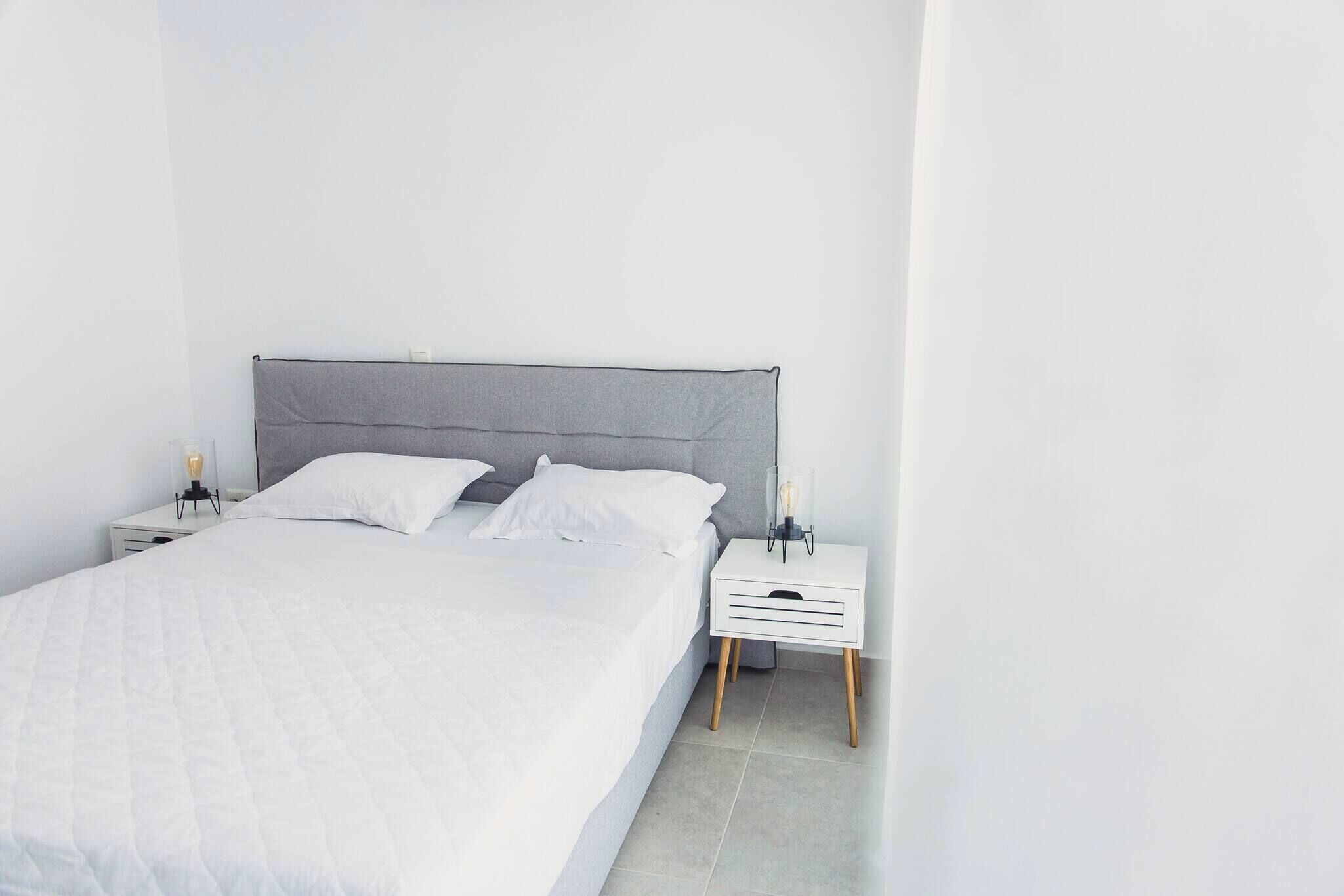 A bed with a grey headboard, white bedding, and a bedside table with a lamp.