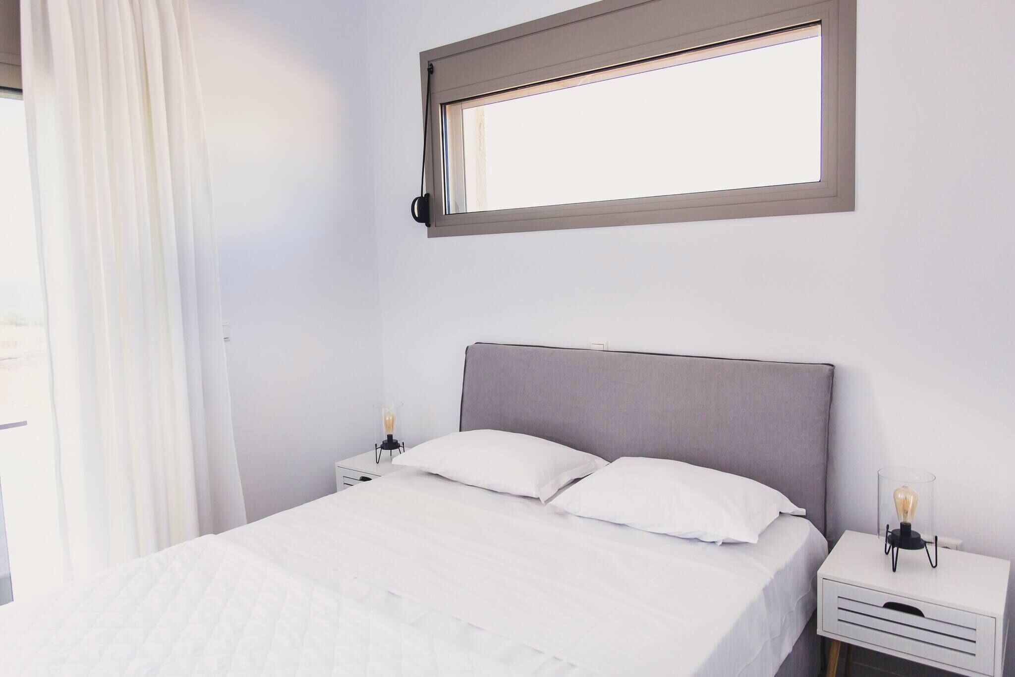A bedroom with a bed, white bedding, a nightstand with a lamp, and a window with a view.