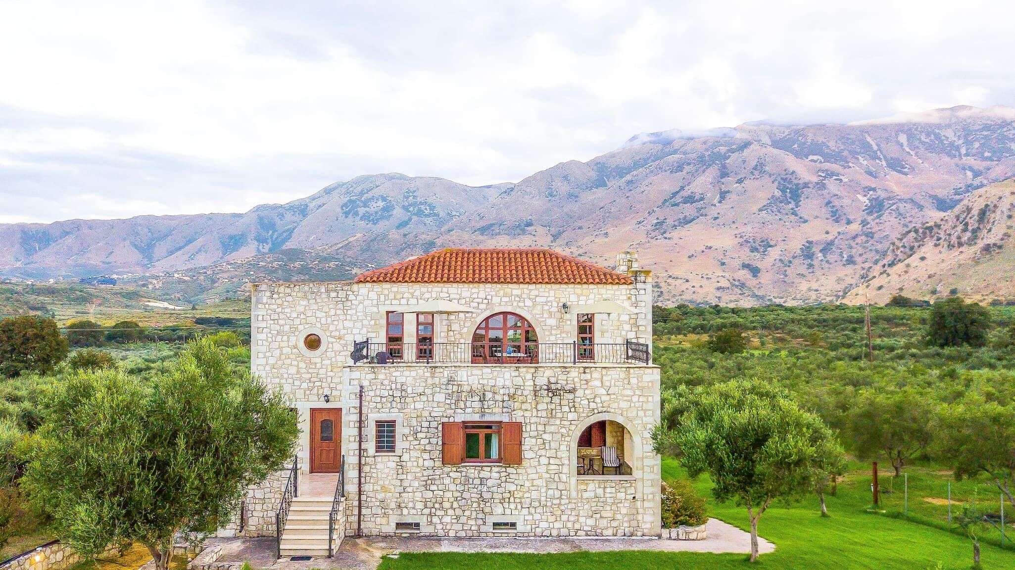 A stone building with a red-tiled roof, arched windows, and a balcony, surrounded by lush greenery and mountains.