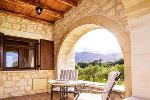A stone-walled room with a wooden ceiling, a window with wooden shutters, a round table with a book, two striped chairs, and a view of greenery and mountains. - Villa Ostria Near Georgioupolis - 5BD 4BA 10pl (Apokoronas)