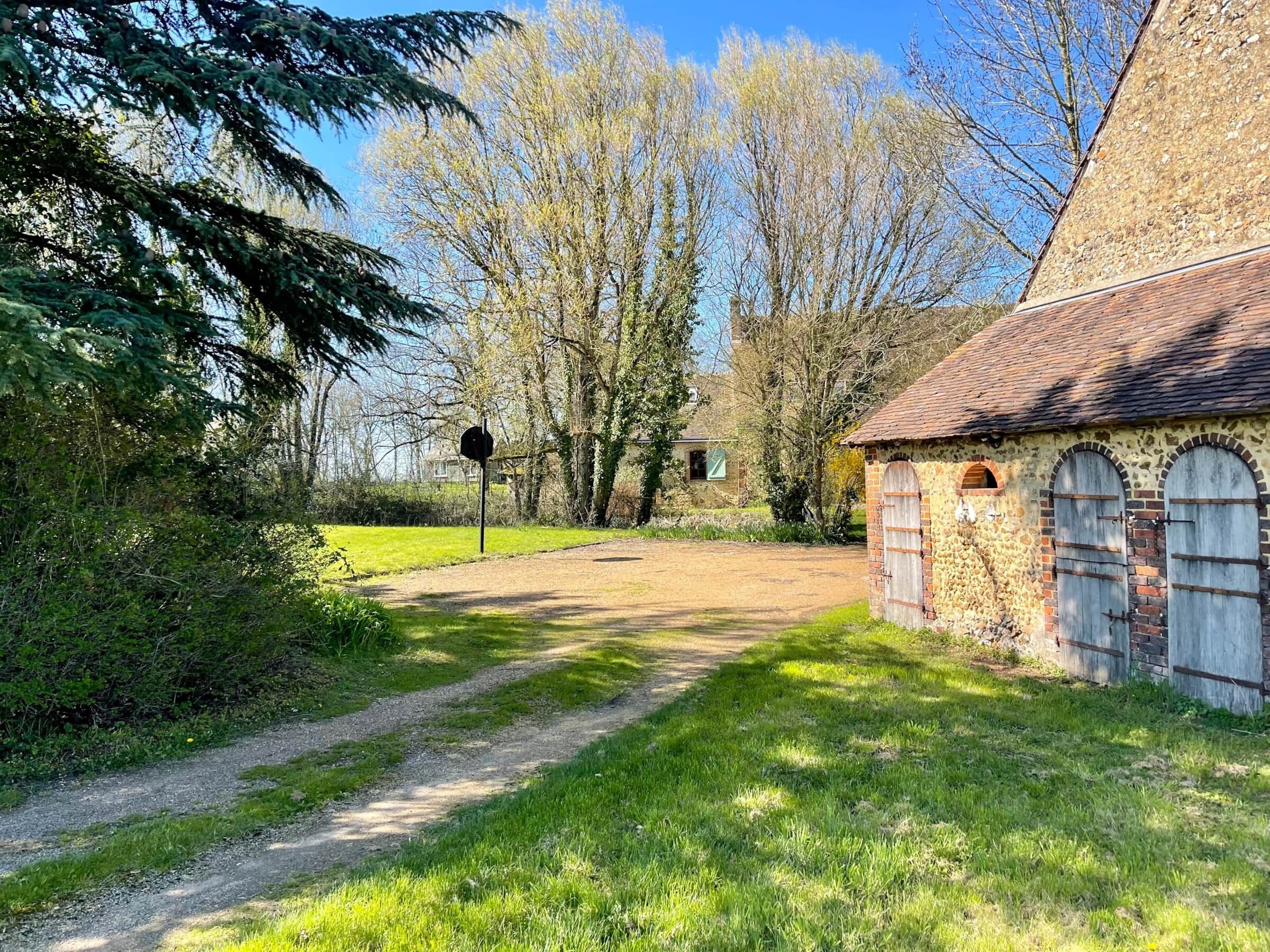 Un sentier rural menant à un bâtiment en pierre doté d’arches au niveau des fenêtres et d’un toit de chaume.
