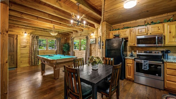 Interior de una cabaña de madera con mesa de billar, comedor y zona de cocina que incluye un refrigerador y estufa.
